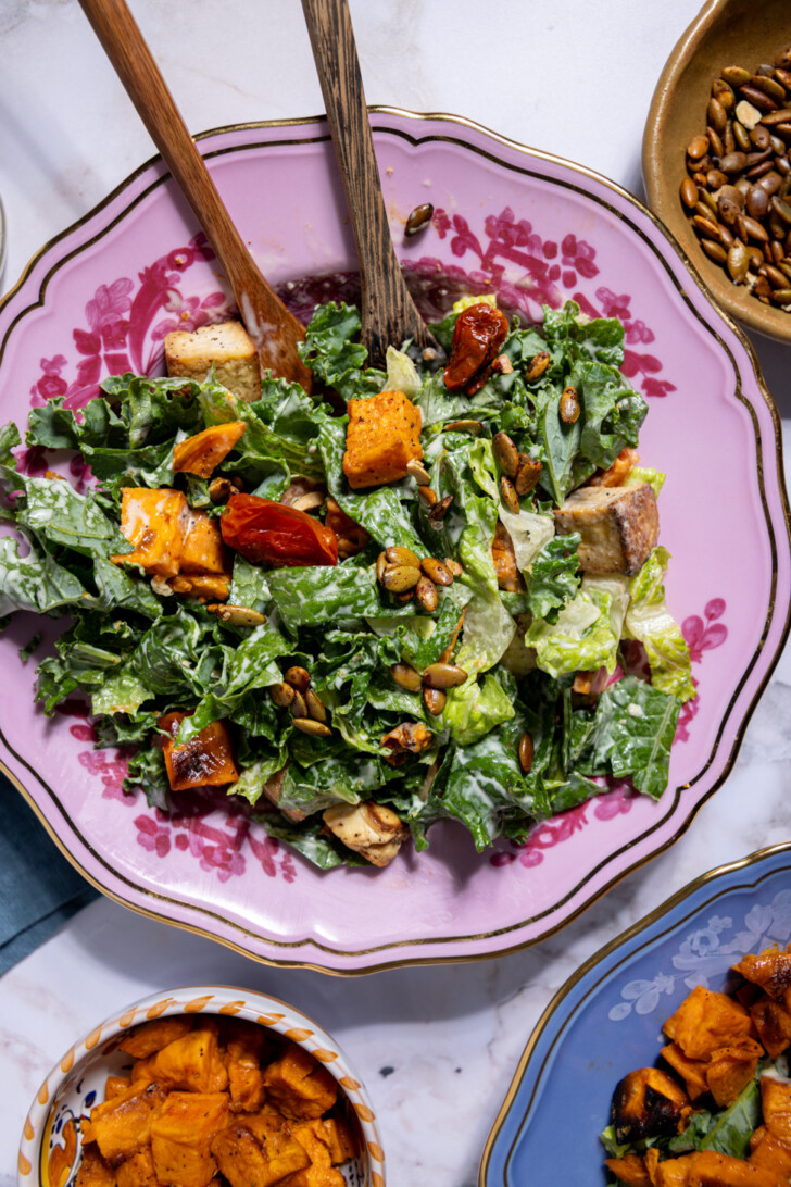 Fall salad recipe featuring crispy tofu, jammy roasted tomatoes, sweet potatoes, and candied pumpkin seeds tossed in vegan Caesar dressing.