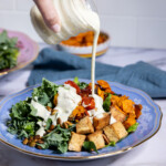 Pouring creamy vegan Caesar dressing over a fall Caesar salad with tofu, sweet potatoes, and roasted tomatoes.
