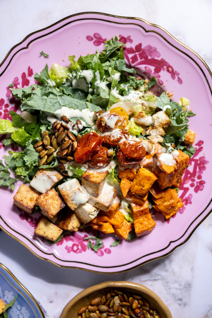 Close-up of a vegan fall salad with crispy tofu, jammy roasted tomatoes, pumpkin seeds, and dairy-free Caesar dressing.