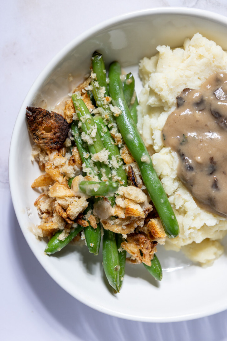Bowl of vegan green bean casserole with crispy topping next to mashed potatoes.